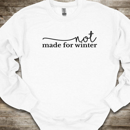 A white crewneck sweatshirt with the phrase 'not made for winter' printed in white across the chest, laid flat on a rustic wooden background.
