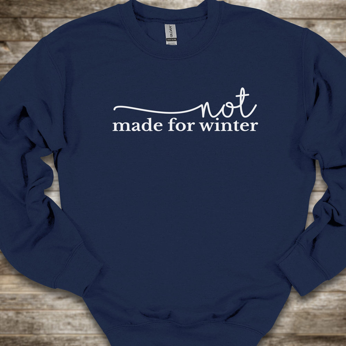 A navy blue crewneck sweatshirt with the phrase 'not made for winter' printed in white across the chest, laid flat on a rustic wooden background.