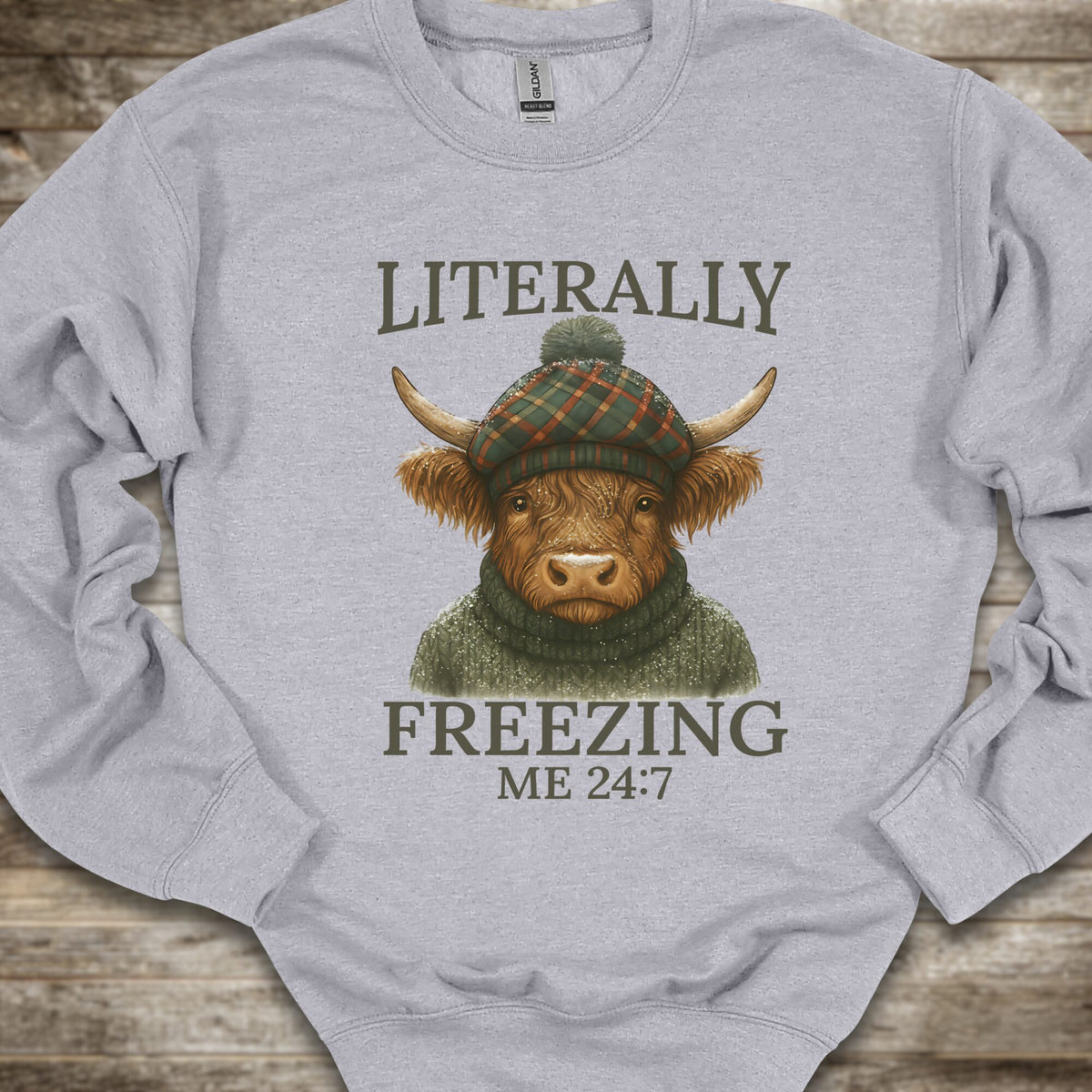 A light gray crewneck sweatshirt laid flat on a rustic wooden surface, featuring a graphic of a horned cow wearing a green plaid beanie and a green knit sweater, with the text 'LITERALLY FREEZING ME 24:7' printed in dark olive lettering.