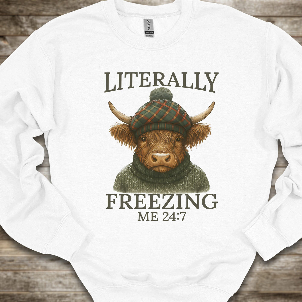 A white long-sleeve crewneck sweatshirt laid flat on a rustic wooden background, featuring a graphic of a highland cow wearing a green plaid beanie and a green sweater, with the text 'LITERALLY FREEZING ME 24:7' printed in dark gray.