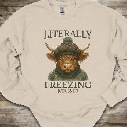 A cream-colored crewneck sweatshirt laid flat on a rustic wooden background, featuring a graphic of a highland cow in a green plaid beanie and green sweater with the text 'LITERALLY FREEZING ME 24:7'.