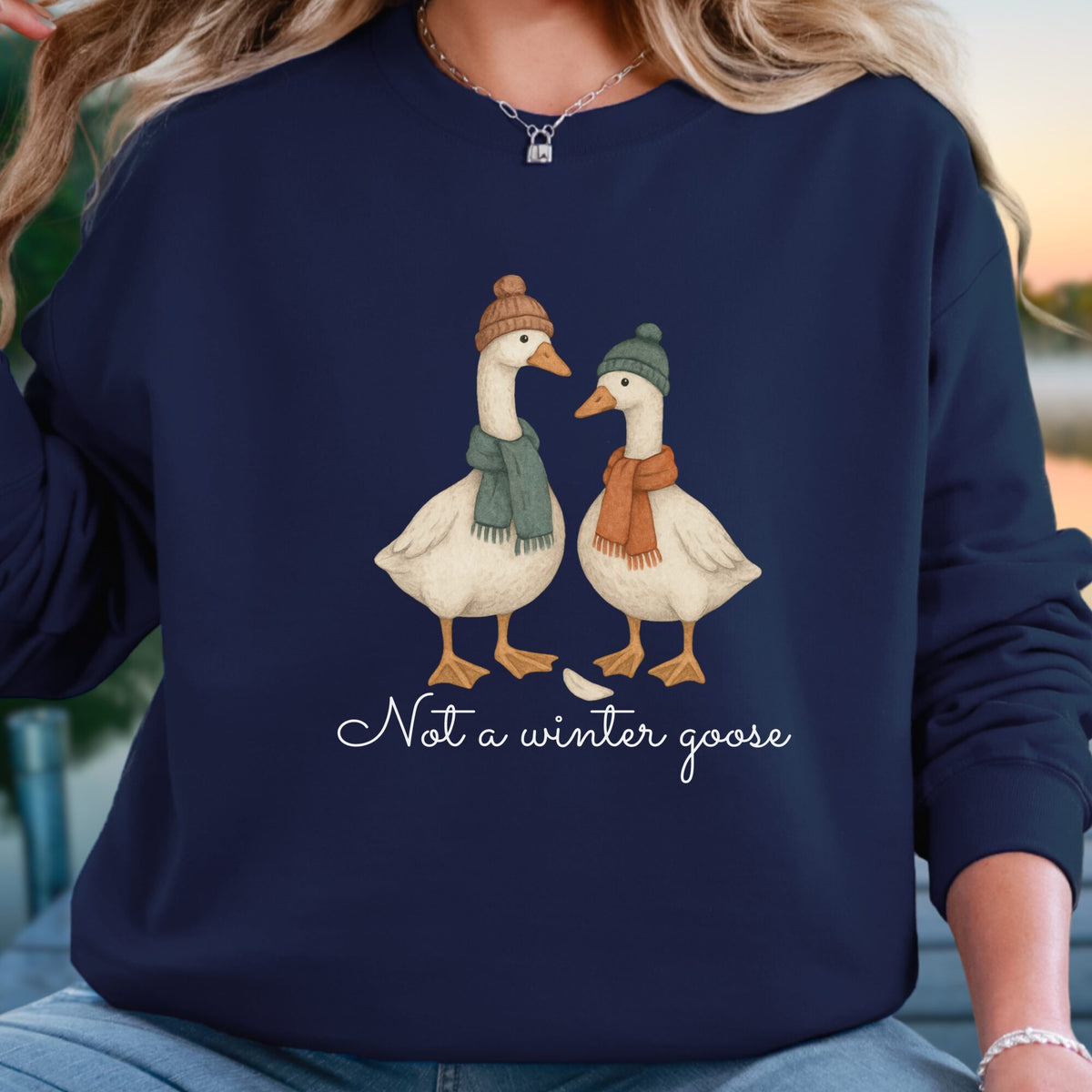 A woman wearing a navy blue sweatshirt with a graphic of two cartoon geese facing each other, one in a green hat and scarf and one in an orange hat and scarf, with the text 'Not a winter goose' below. The background is a softly blurred outdoor setting.