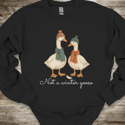 A black crewneck sweatshirt featuring a graphic of two white geese wearing winter hats and scarves, with the text 'Not a winter goose' printed below in white script, laid flat on a rustic wooden background.