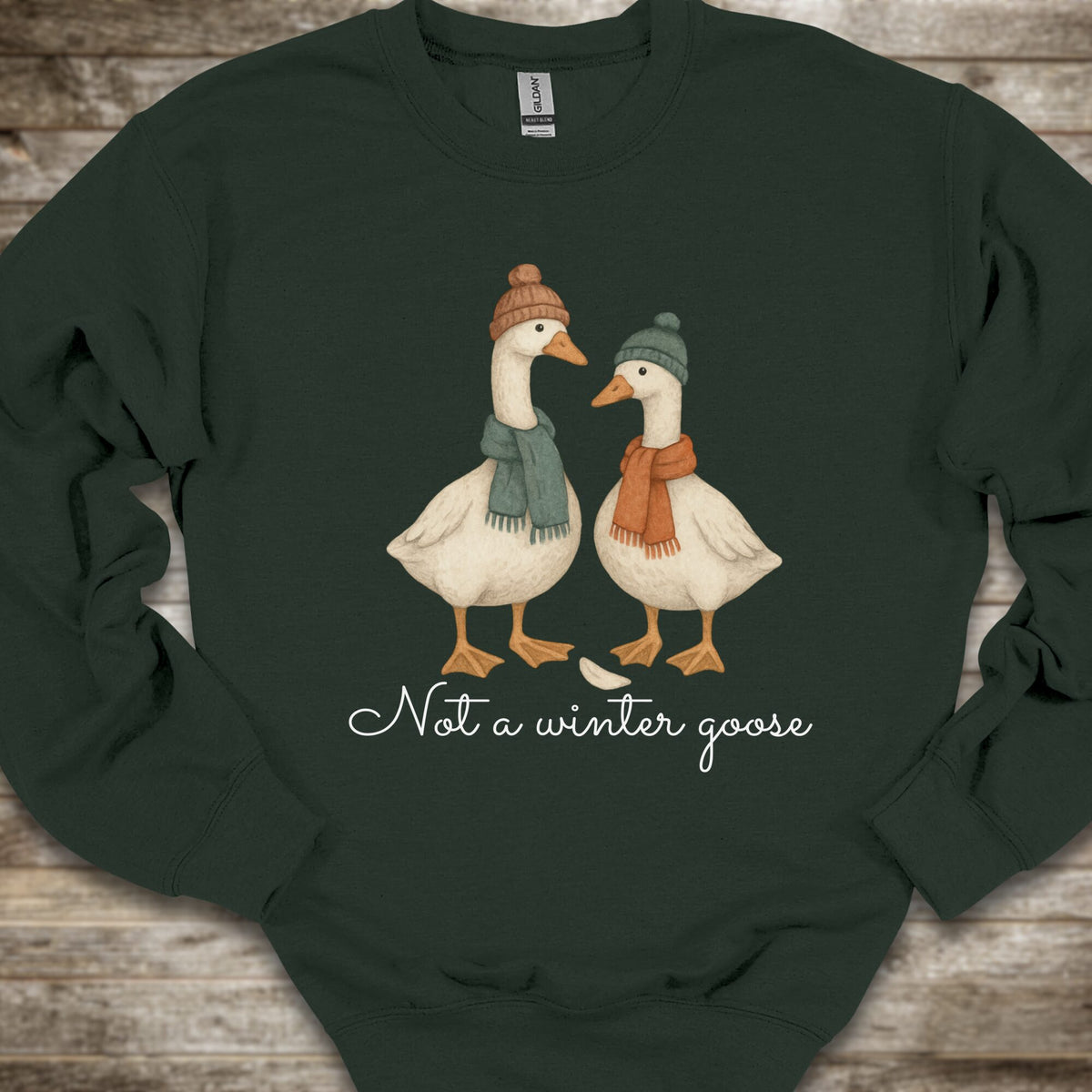 A dark forest green crewneck sweatshirt laid flat on a rustic wooden background. The front of the sweatshirt features a graphic of two white geese wearing winter hats and scarves, with the text 'Not a winter goose' written in white script underneath.