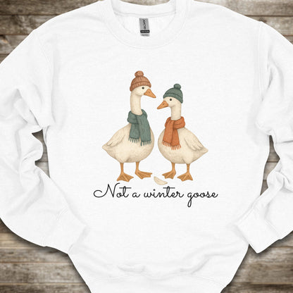 A white crewneck sweatshirt laid flat on a rustic wooden background, featuring a graphic of two geese wearing winter hats and scarves with the text 'Not a winter goose' printed below.