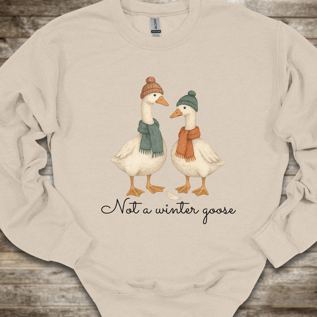 A beige crewneck sweatshirt laid flat on a rustic wooden surface, featuring a graphic of two white geese wearing winter hats and scarves with the text 'Not a winter goose' printed below.