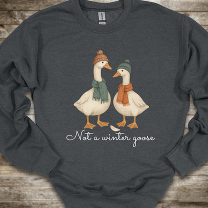A dark gray heather crewneck sweatshirt laid flat on a rustic wooden background, featuring a graphic of two white winter geese wearing orange and green scarves with the text 'Not a winter goose' printed below.