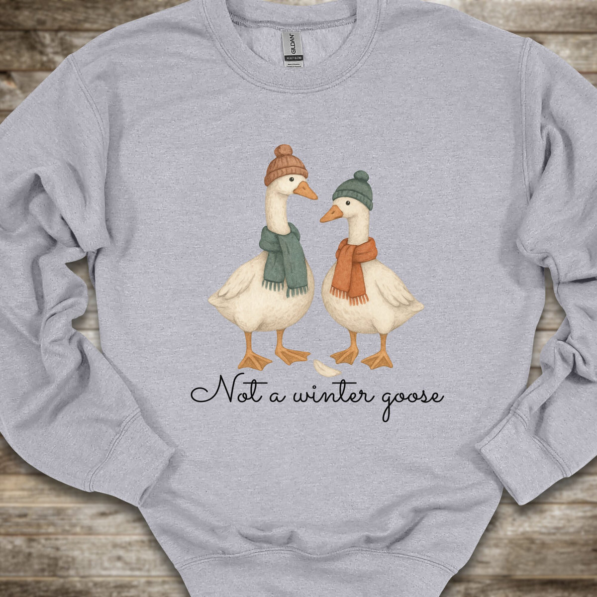 A light heather gray long-sleeve sweatshirt laid flat on a rustic wood background, featuring a graphic of two white geese wearing knit beanies and scarves, with the text 'Not a winter goose' printed below.