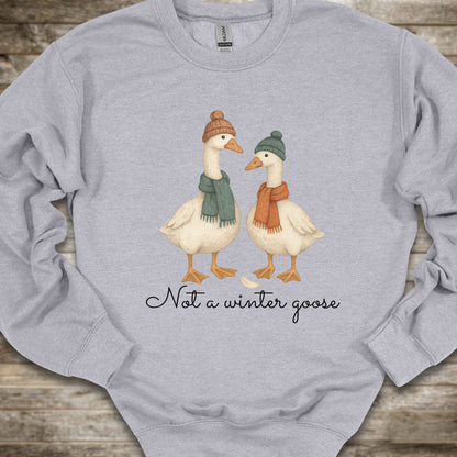 A light heather gray long-sleeve sweatshirt laid flat on a rustic wood background, featuring a graphic of two white geese wearing knit beanies and scarves, with the text 'Not a winter goose' printed below.