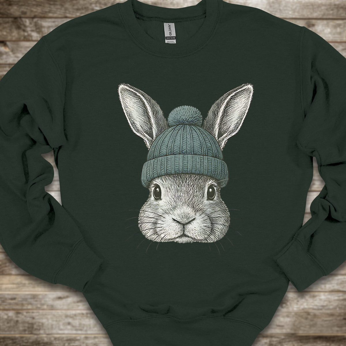 A dark forest green crewneck sweatshirt laid flat on a rustic wooden background, featuring a detailed graphic of a gray rabbit wearing a teal knit beanie.