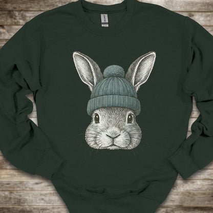 A dark forest green crewneck sweatshirt laid flat on a rustic wooden background, featuring a detailed graphic of a gray rabbit wearing a teal knit beanie.
