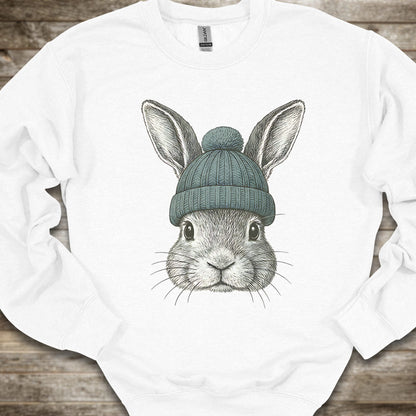 A white crewneck sweatshirt featuring a detailed graphic of a gray rabbit wearing a teal knit beanie, laid flat on a rustic wooden background.