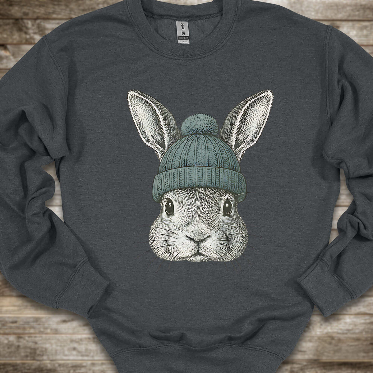A dark charcoal gray crewneck sweatshirt featuring a detailed graphic of a gray rabbit wearing a teal knit beanie, laid flat on a rustic wooden background.