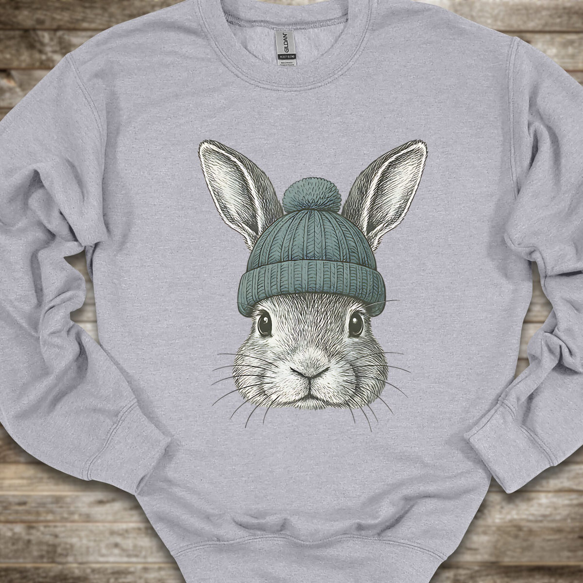 A light gray crewneck sweatshirt featuring a detailed graphic of a gray rabbit wearing a teal knit beanie, laid flat on a rustic wooden background.