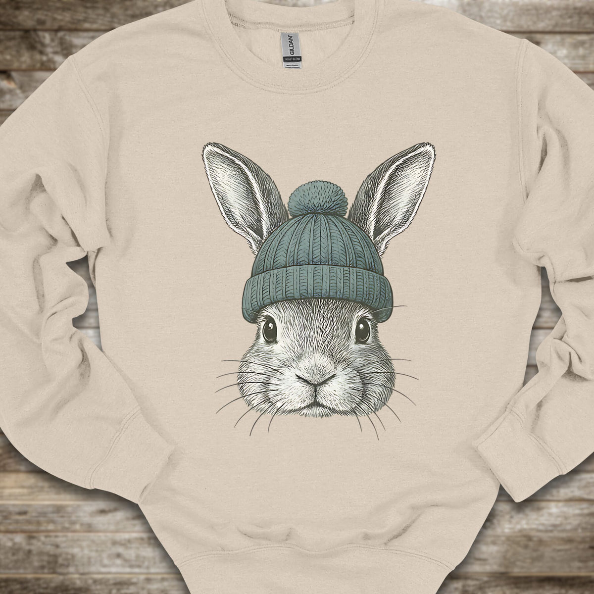 A beige crewneck sweatshirt featuring a detailed graphic of a gray rabbit wearing a teal knit beanie, laid flat on a rustic wooden background.