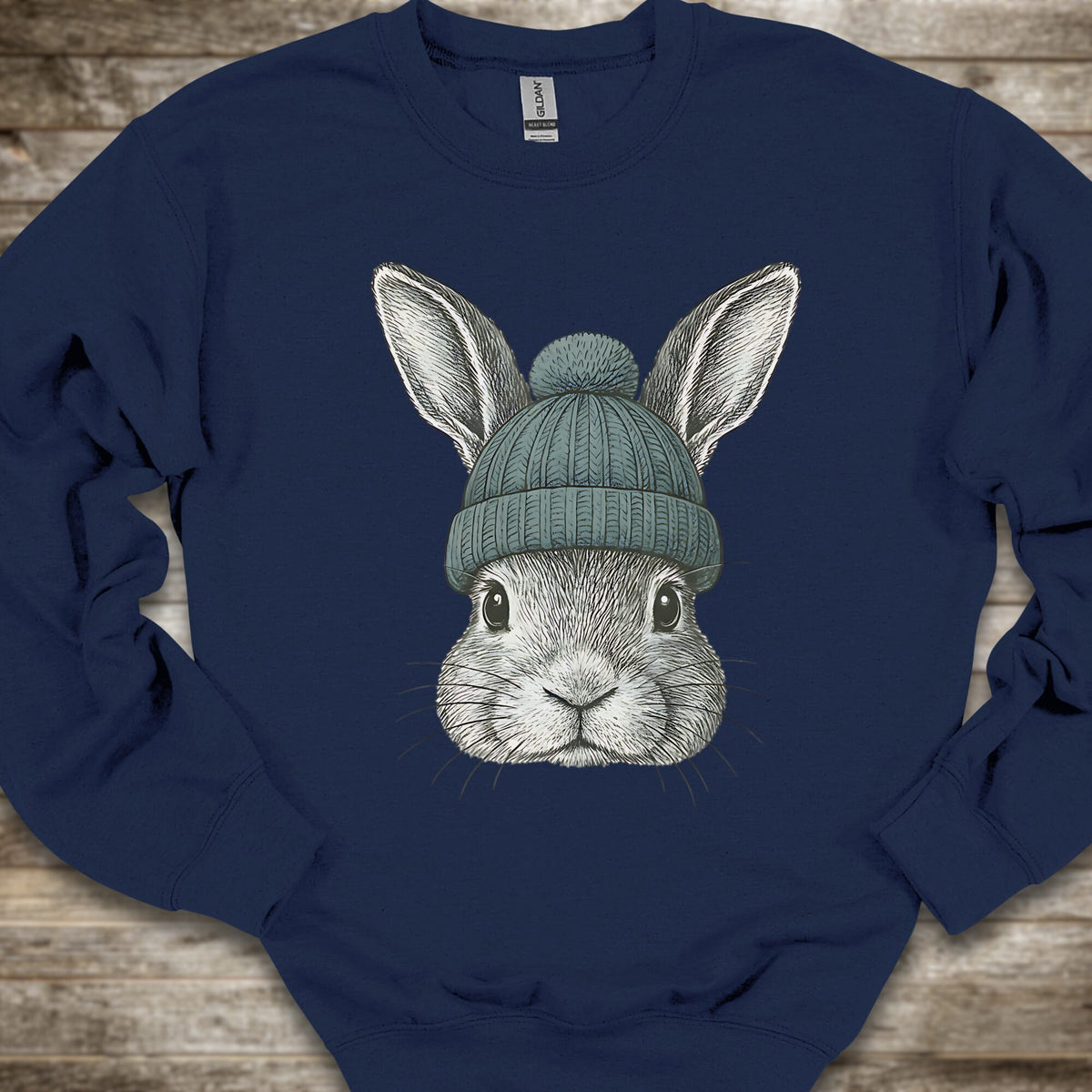A navy blue crewneck sweatshirt featuring a detailed graphic of a gray rabbit wearing a teal knit beanie, laid flat on a rustic wooden background.