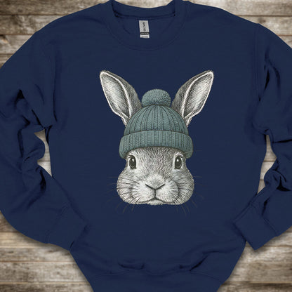A navy blue crewneck sweatshirt featuring a detailed graphic of a gray rabbit wearing a teal knit beanie, laid flat on a rustic wooden background.