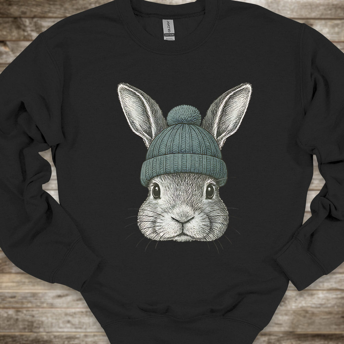 A black crewneck sweatshirt featuring a detailed graphic of a gray rabbit wearing a teal knit beanie, laid flat on a rustic wooden background.