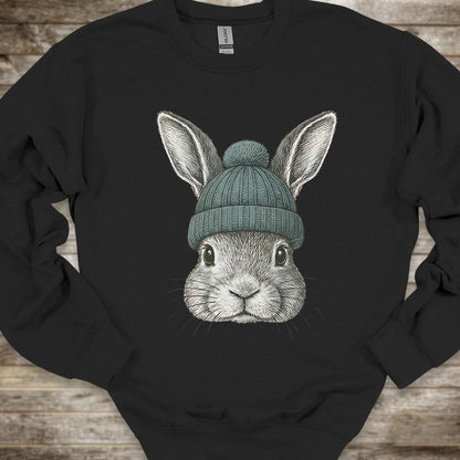 A black crewneck sweatshirt featuring a detailed graphic of a gray rabbit wearing a teal knit beanie, laid flat on a rustic wooden background.
