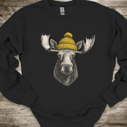 A black crewneck sweatshirt featuring a graphic print of a moose head wearing a yellow knit beanie, laid flat on a rustic wooden background.