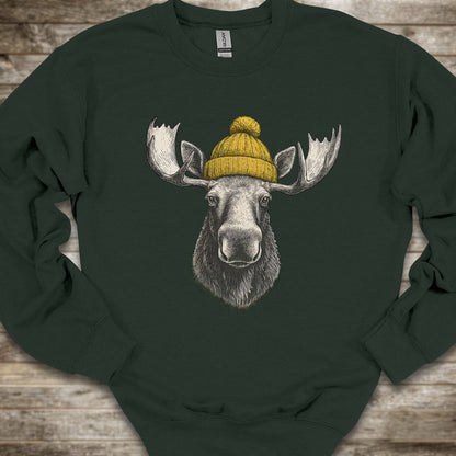 A dark forest green crewneck sweatshirt featuring a detailed graphic of a moose head with large antlers wearing a yellow knit beanie, laid flat on a rustic wooden background.