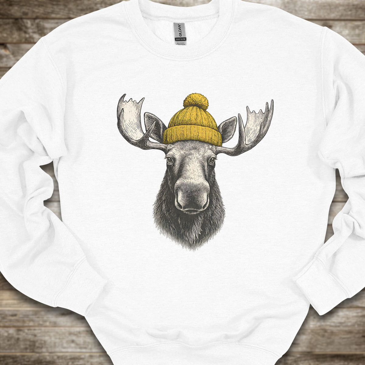 A white crewneck sweatshirt featuring a graphic print of a moose head with large antlers wearing a yellow knit beanie, laid flat on a rustic wooden background.