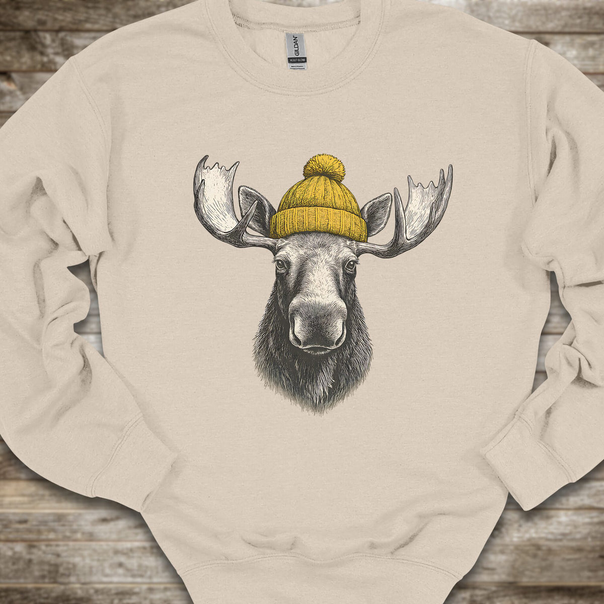 A beige crewneck sweatshirt featuring a graphic print of a moose head with large antlers wearing a yellow knit beanie, laid flat on a rustic wooden surface.