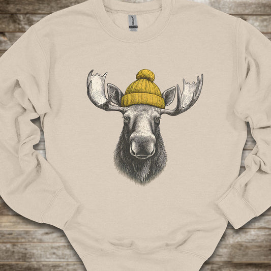 A beige crewneck sweatshirt featuring a graphic print of a moose head with large antlers wearing a yellow knit beanie, laid flat on a rustic wooden surface.