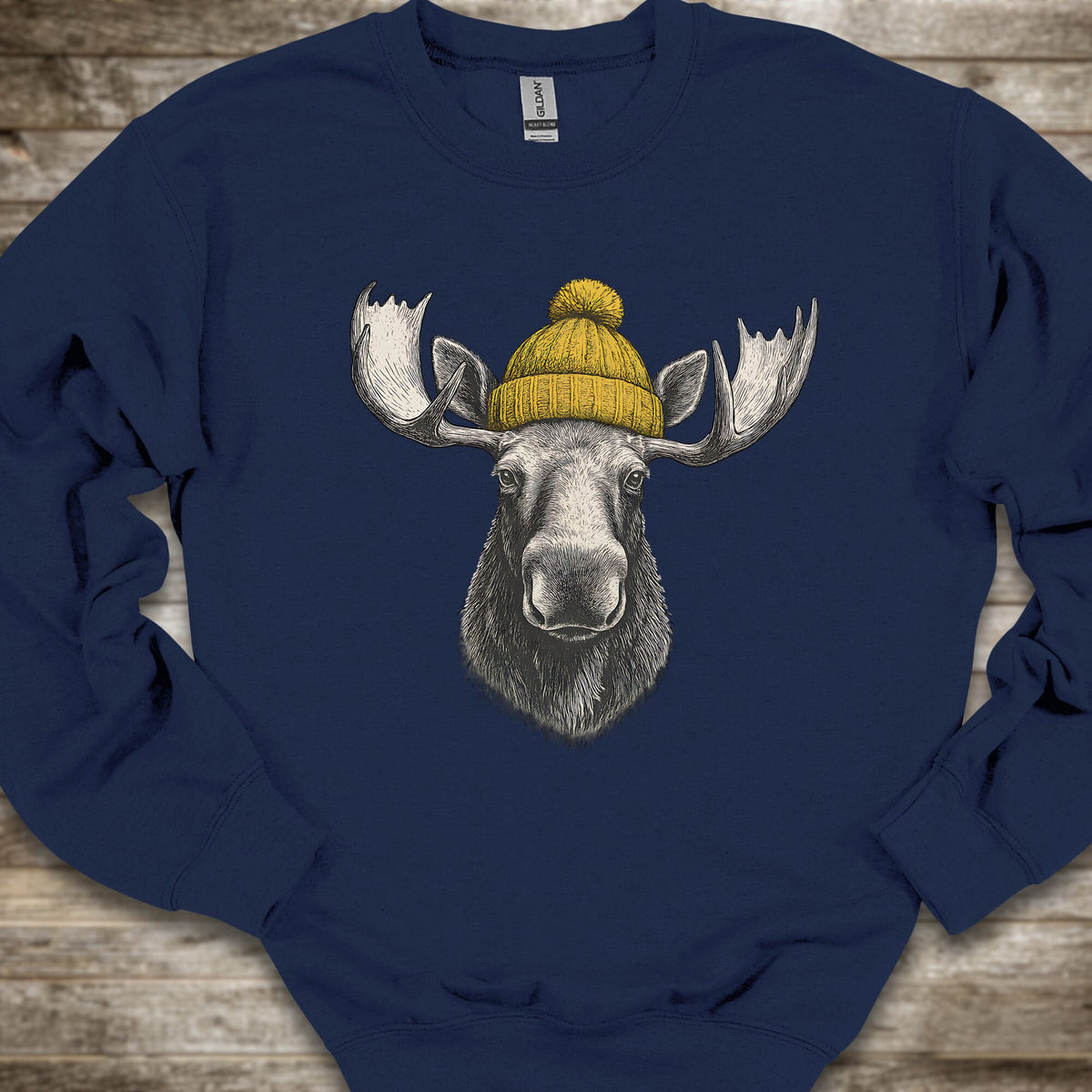 A navy blue crewneck sweatshirt laid flat on a rustic wooden background, featuring a graphic print of a moose head wearing a yellow knit beanie.