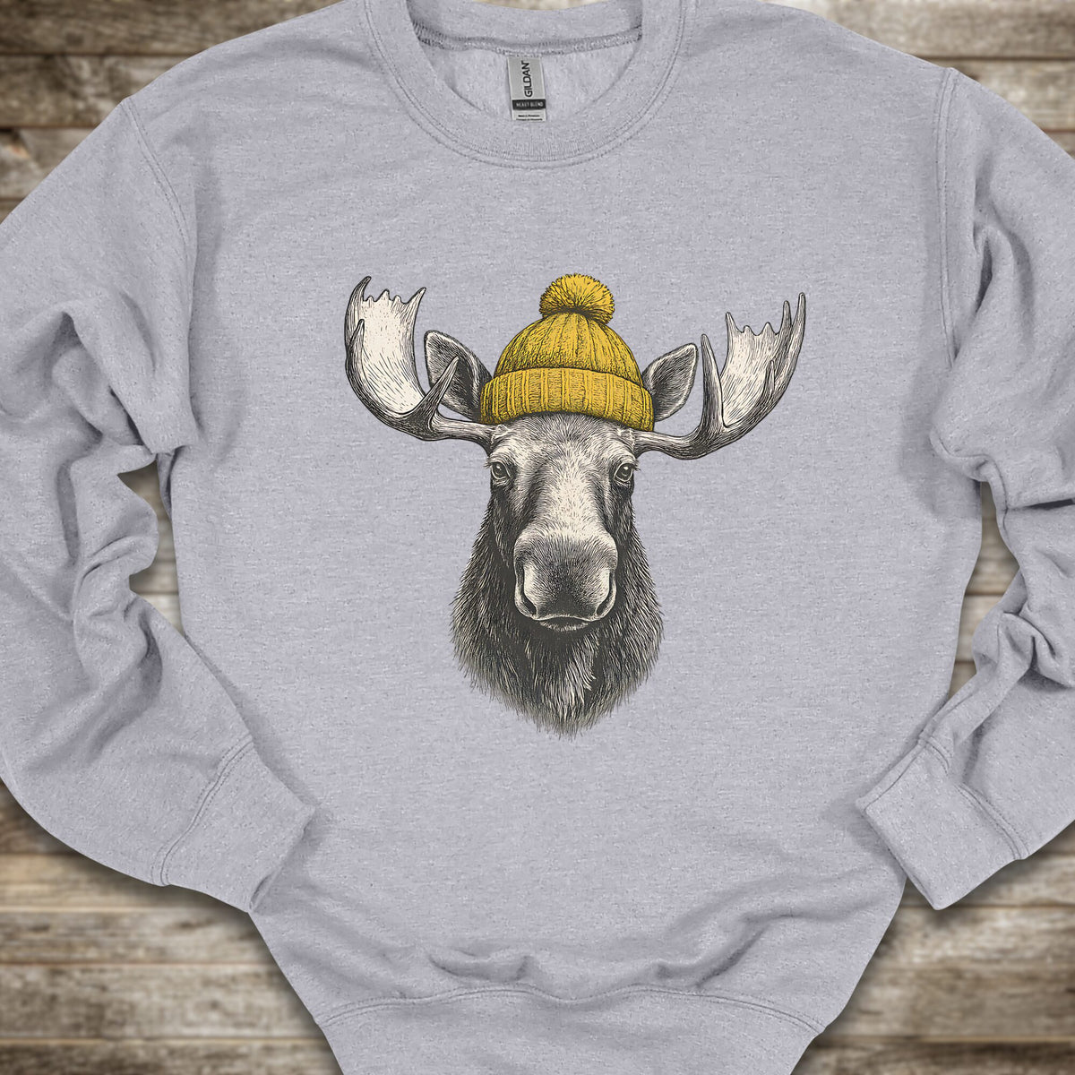 A light gray crewneck sweatshirt featuring a graphic print of a moose head wearing a yellow knit beanie, laid flat on a rustic wooden surface.