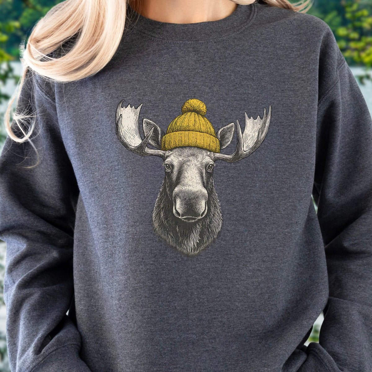 A person wearing a dark heather gray crewneck sweatshirt with a graphic of a moose head wearing a yellow knit beanie. The background is a soft-focus outdoor green foliage.