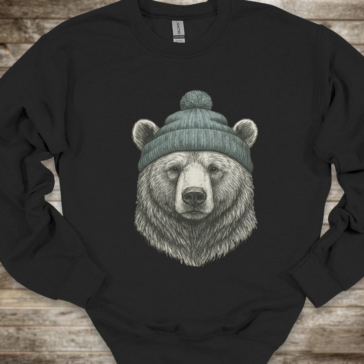 A black crewneck sweatshirt featuring a detailed graphic of a bear's head wearing a green knit beanie, laid flat on a rustic wooden background.