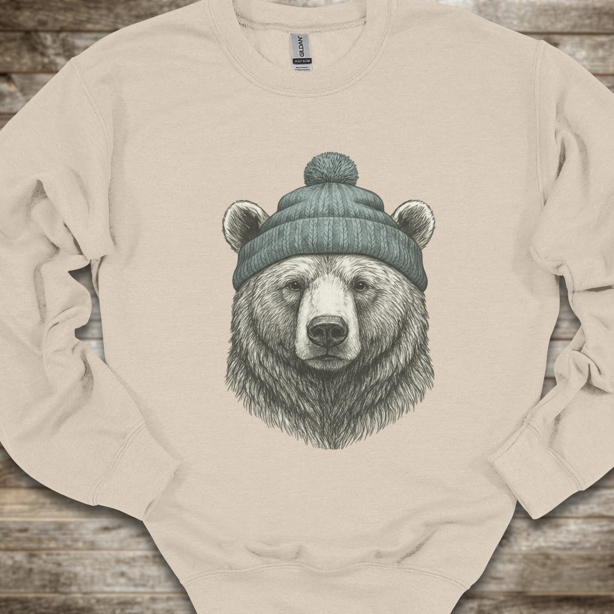 A sand crewneck sweatshirt featuring a detailed graphic of a bear's head wearing a green knit beanie, laid flat on a rustic wooden background.