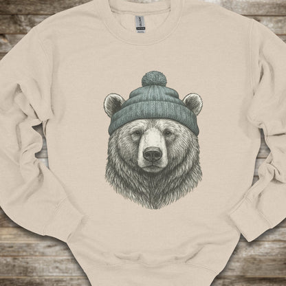 A sand crewneck sweatshirt featuring a detailed graphic of a bear's head wearing a green knit beanie, laid flat on a rustic wooden background.