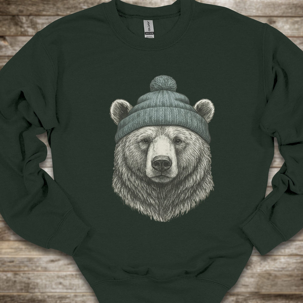 A Forest Green crewneck sweatshirt featuring a detailed graphic of a bear's head wearing a green knit beanie, laid flat on a rustic wooden background.