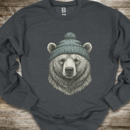 A Dark Heather crewneck sweatshirt featuring a detailed graphic of a bear's head wearing a green knit beanie, laid flat on a rustic wooden background.