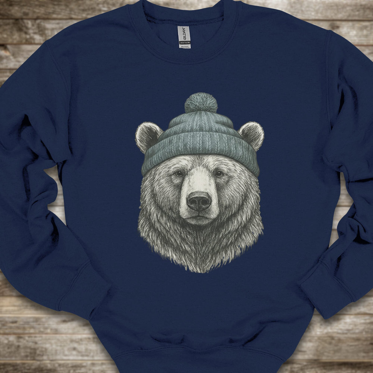 A navy crewneck sweatshirt featuring a detailed graphic of a bear's head wearing a green knit beanie, laid flat on a rustic wooden background.