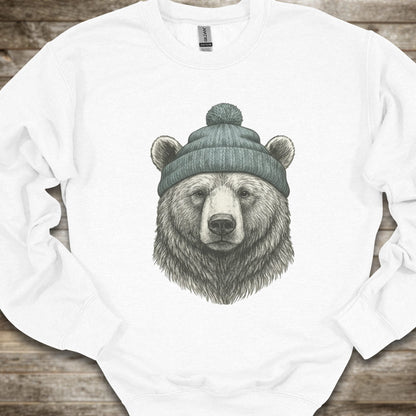 A white crewneck sweatshirt featuring a detailed graphic of a bear's head wearing a green knit beanie, laid flat on a rustic wooden background.