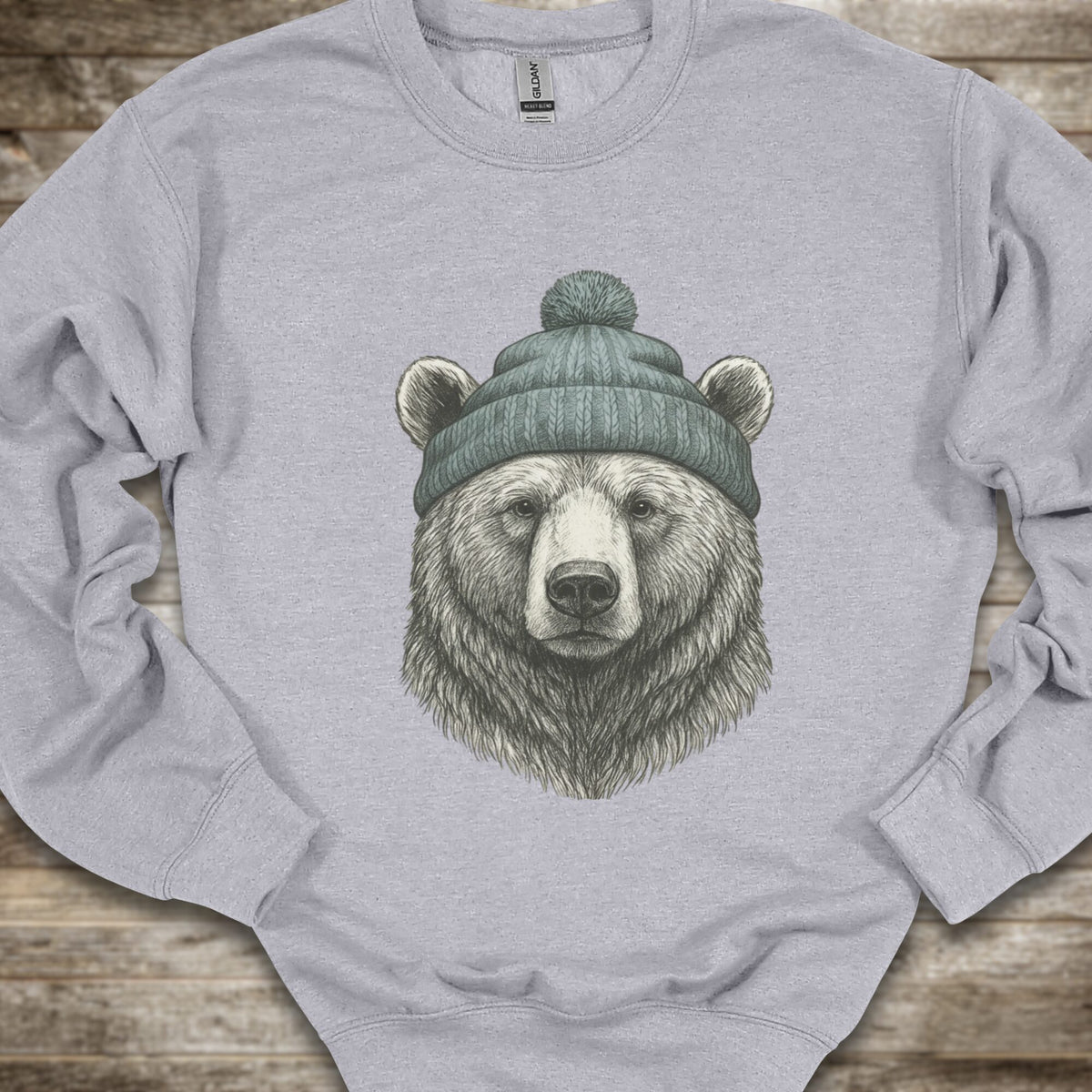 A light gray crewneck sweatshirt featuring a detailed graphic of a bear's head wearing a green knit beanie, laid flat on a rustic wooden background.