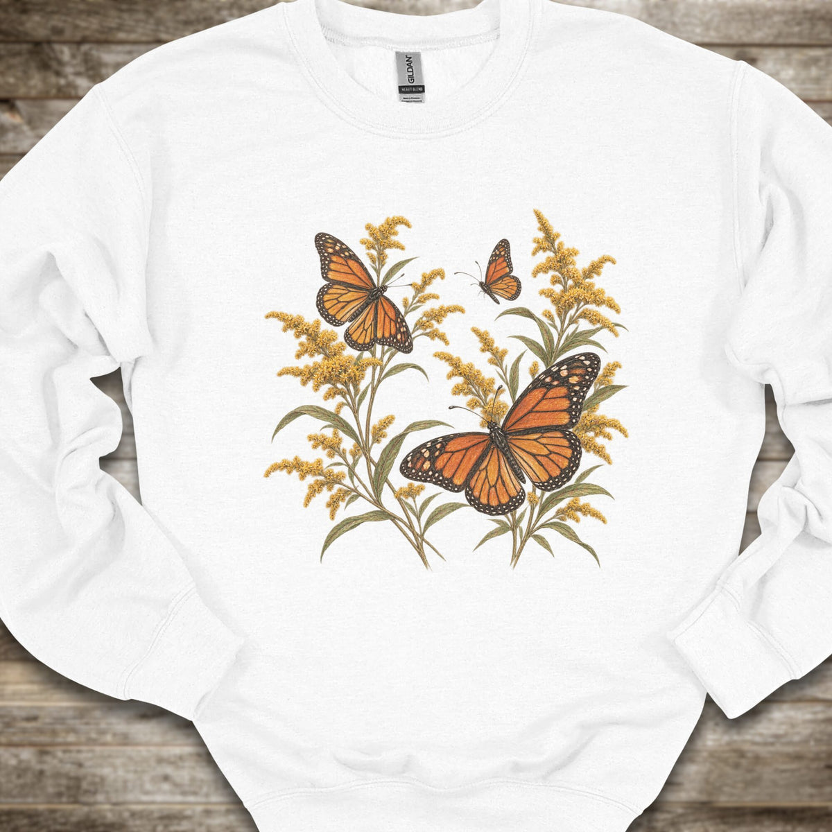 A white crewneck sweatshirt featuring a graphic print of monarch butterflies and golden flowers, laid flat on a rustic wooden background.