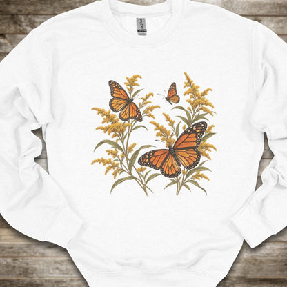 A white crewneck sweatshirt featuring a graphic print of monarch butterflies and golden flowers, laid flat on a rustic wooden background.