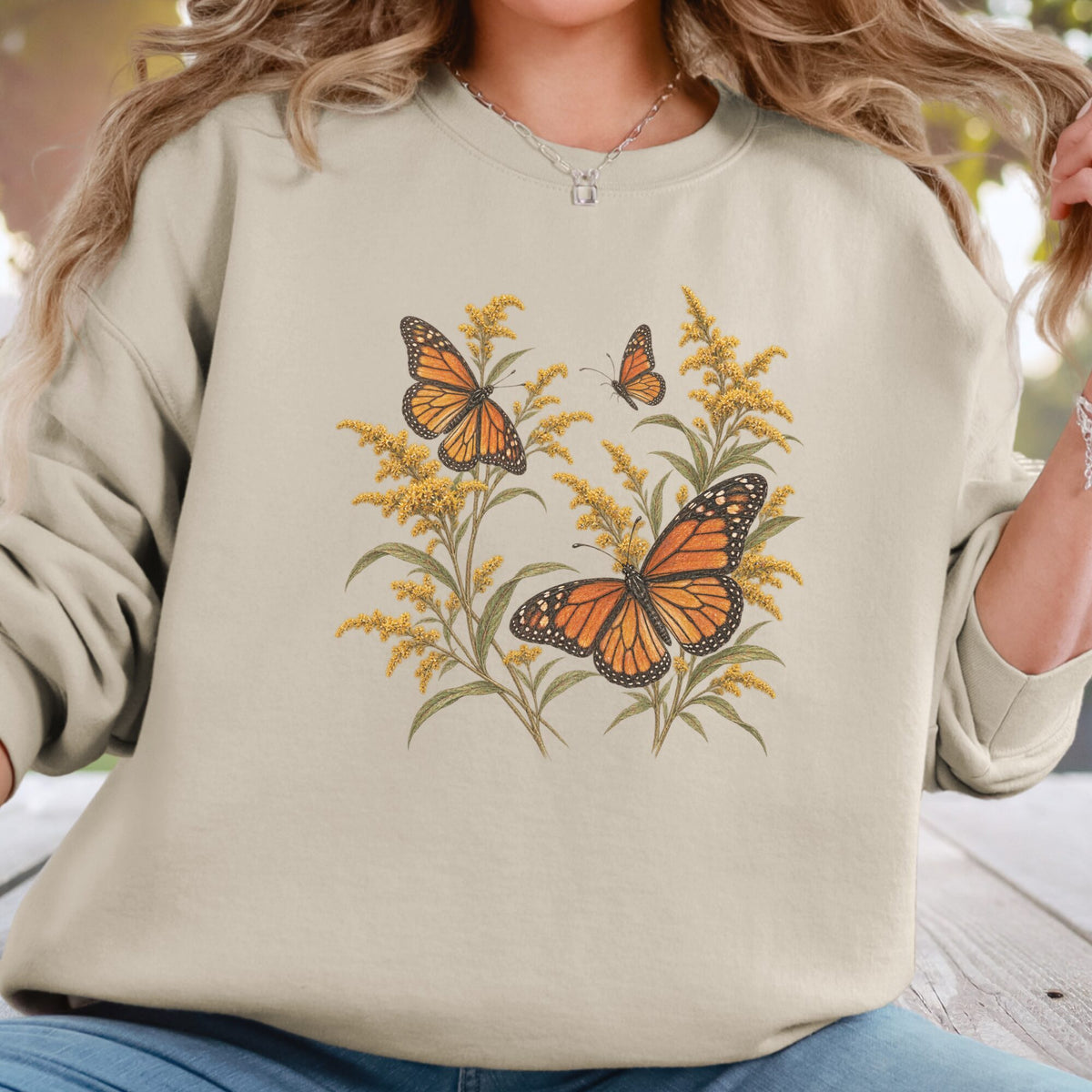 A woman wearing a beige crewneck sweatshirt with a graphic print of orange monarch butterflies and yellow golden flowers. The photo is cropped from the shoulders to the hips, set against a softly blurred outdoor background.