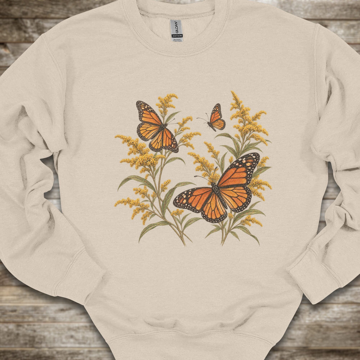 A cream-colored crewneck sweatshirt featuring a graphic print of two orange monarch butterflies perched on yellow wildflowers, laid flat on a rustic wooden surface.