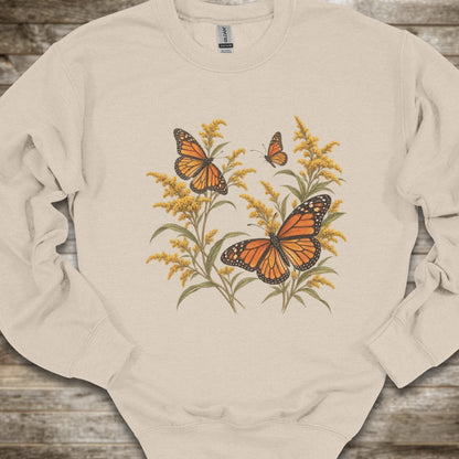 A cream-colored crewneck sweatshirt featuring a graphic print of two orange monarch butterflies perched on yellow wildflowers, laid flat on a rustic wooden surface.