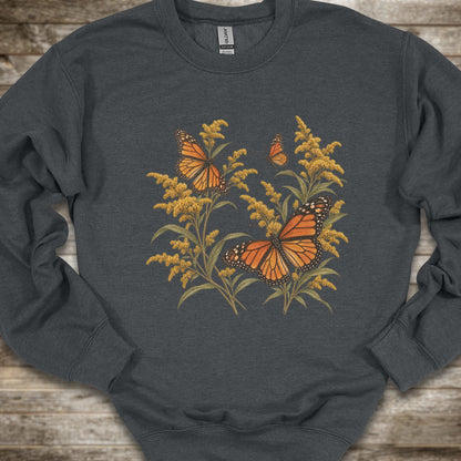 A dark gray crewneck sweatshirt featuring a graphic print of monarch butterflies and golden flowers, laid flat on a rustic wooden background.