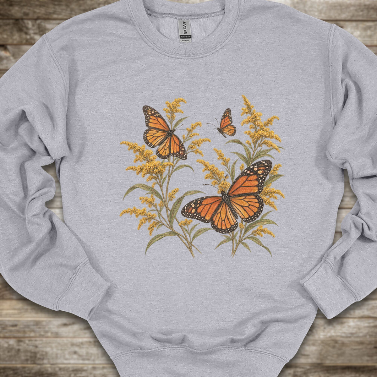 A light heather gray crewneck sweatshirt featuring a detailed graphic print of monarch butterflies and golden yellow wildflowers, laid flat on a rustic wooden surface.