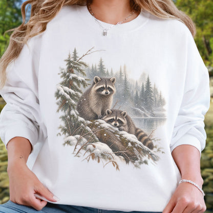 A person wearing a white crewneck sweatshirt with a detailed graphic print of two raccoons in a snowy pine forest. The background is a soft-focus outdoor green setting.