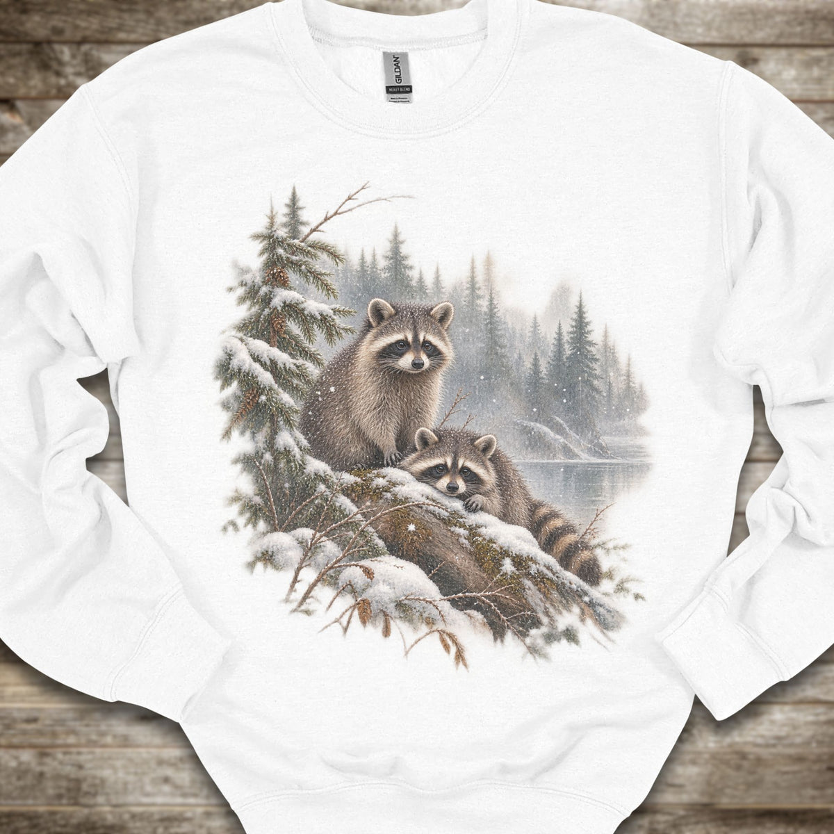A white crewneck sweatshirt featuring a detailed graphic print of two raccoons in a snowy winter forest scene, laid flat on a rustic wooden background.