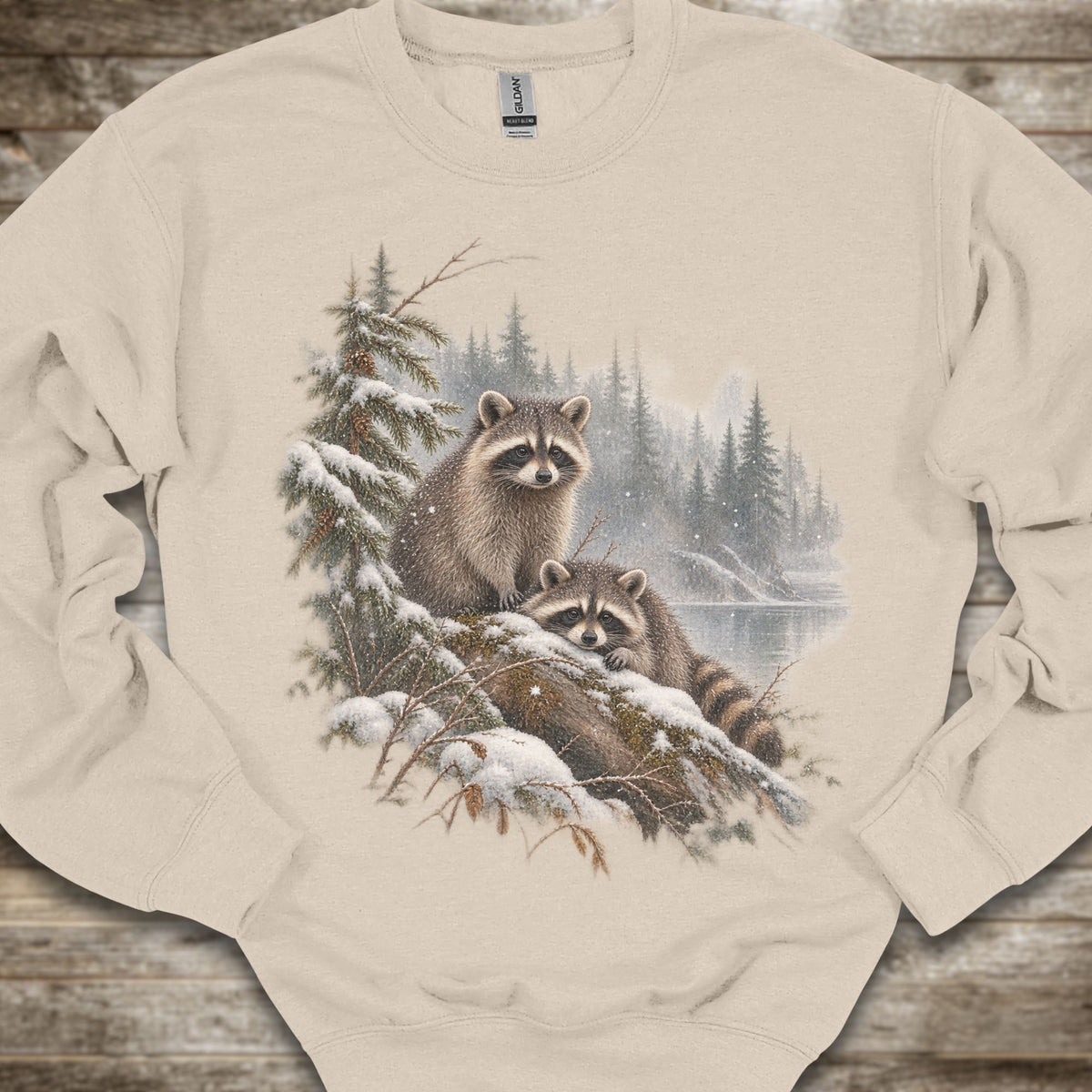 A beige crewneck sweatshirt featuring a detailed graphic print of two raccoons in a snowy pine forest. The sweatshirt is laid flat on a rustic wooden surface.