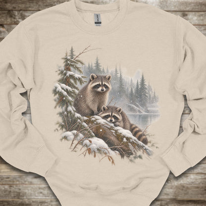 A beige crewneck sweatshirt featuring a detailed graphic print of two raccoons in a snowy pine forest. The sweatshirt is laid flat on a rustic wooden surface.